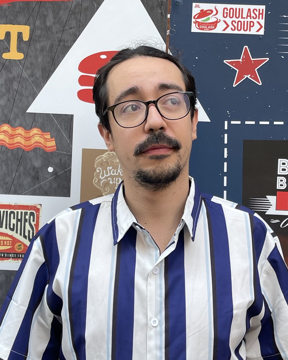 David Figueroa, de óculos e camisa listrada azul e branca, diante de uma parede de cartazes